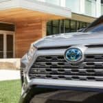 The grille of a silver 2020 Toyota RAV4 Hybrid is shown with a modern building behind it.