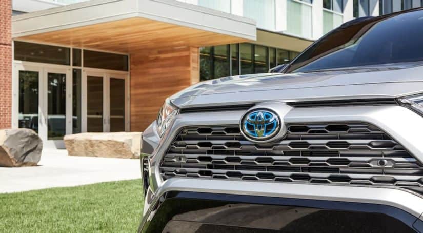 The grille of a silver 2020 Toyota RAV4 Hybrid is shown with a modern building behind it.