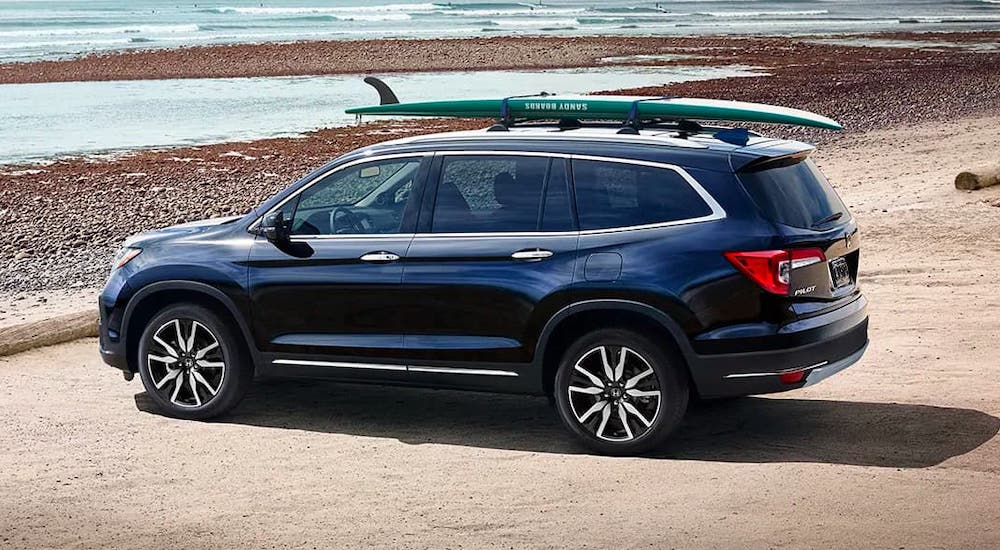 A blue 2020 Honda Pilot Elite with a surfboard on the roof is parked at a beach.