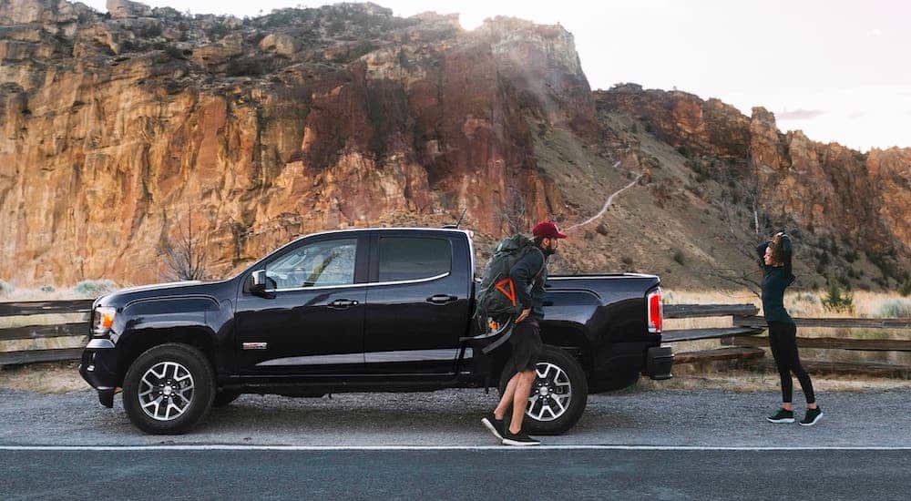 Two hikers are standing next to a black 2020 GMC Canyon All Terrain which is parked in front of mountains.