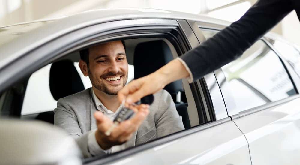 A sales person is handing keys to a man in a silver car.