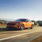 An orange 2018 Ford Mustang is driving on a winding mountain road.