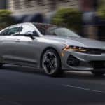 A grey 2021 Kia K5 is driving on a downtown street.