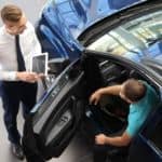A car salesman is talking to a buyer about Certified Pre Owned cars while he sits in a blue vehicle.
