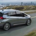 A silver 2020 Chevy Bolt EV, one of the electric Chevy cars, is driving with a city in the distance.