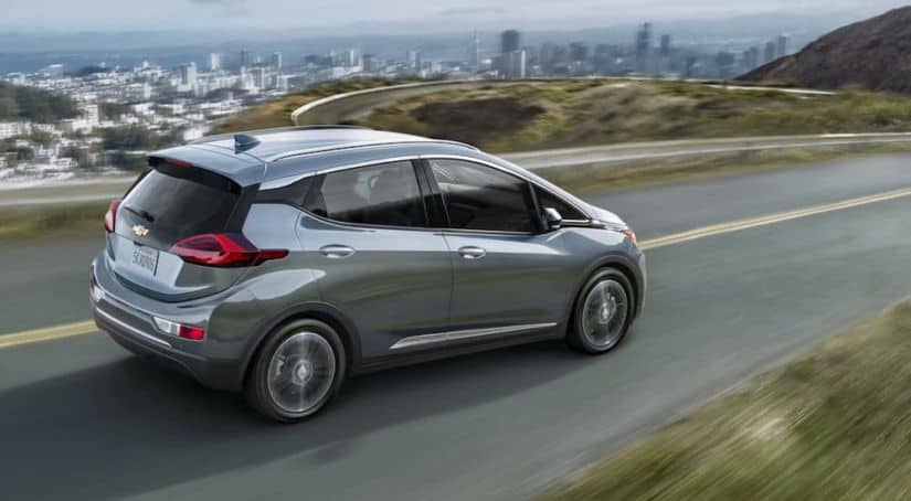 A silver 2020 Chevy Bolt EV, one of the electric Chevy cars, is driving with a city in the distance.
