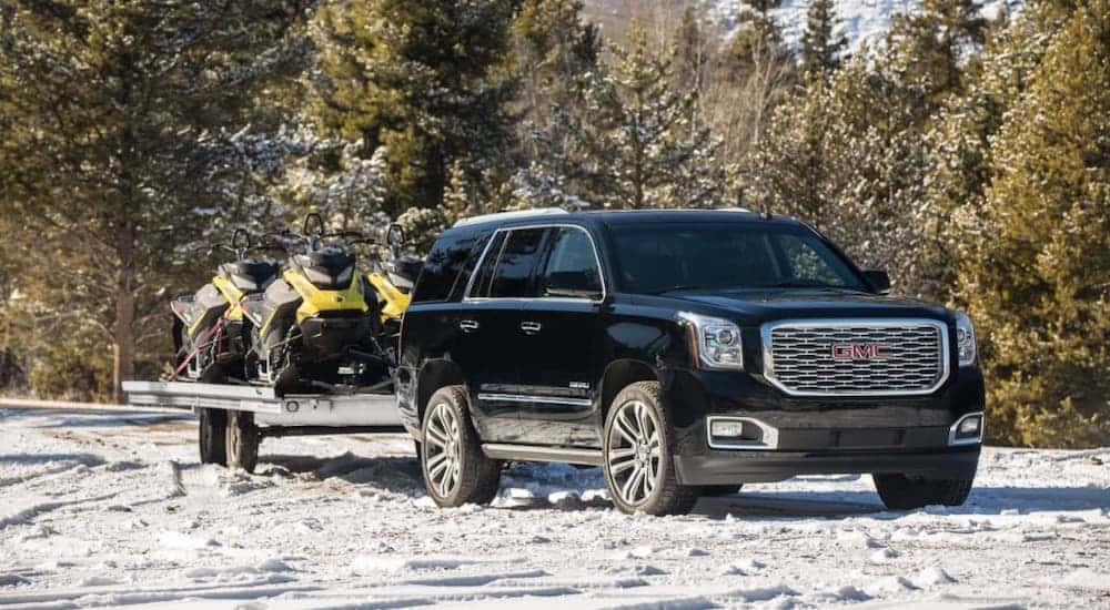 A black 2019 GMC Yukon Denali from a used car dealership is towing snowmobiles on a snowy trail.
