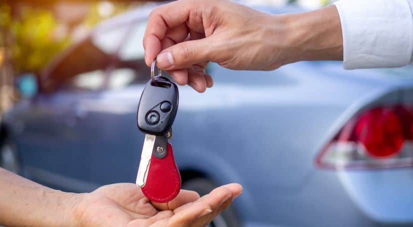 A salesperson is handing keys to someone in front of a used blue car.