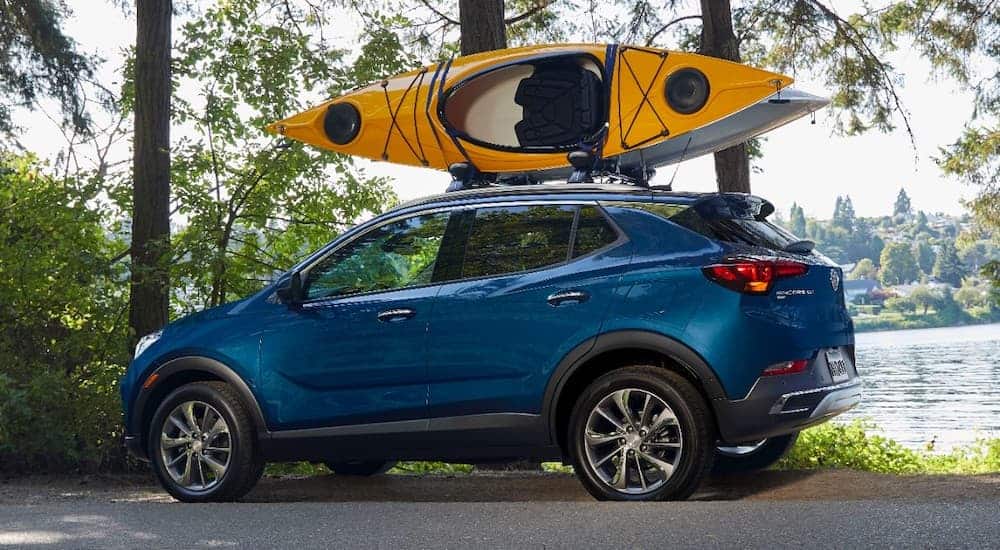 A blue 2020 Buick Encore GX is parked in front of trees and a pond with a kayak on the roof.