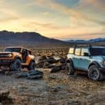 A yellow 2021 Ford Bronco 2-door and a blue Bronco 4-door are parked on rocks in front of a desert and mountains.