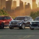 Three Chevy SUVs, a red 2020 Blazer, grey Equinox, and grey Trax, are parked in front of a city at sunset.