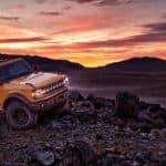 An orange 2021 Ford Bronco 2-door, a new Ford SUV, is parked on rocks in front of a vibrant sunset.