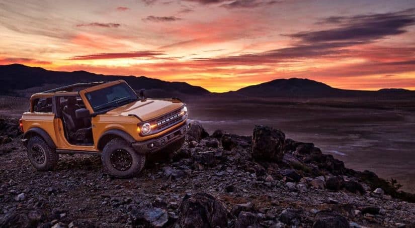An orange 2021 Ford Bronco 2-door, a new Ford SUV, is parked on rocks in front of a vibrant sunset.
