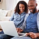 A couple is sitting on the living room floor while looking at an online car dealership on a laptop.