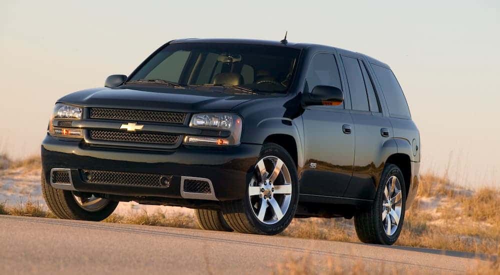An older used car for sale, a black 2009 Chevy Trailblazer SS, is parked in front of a beach.