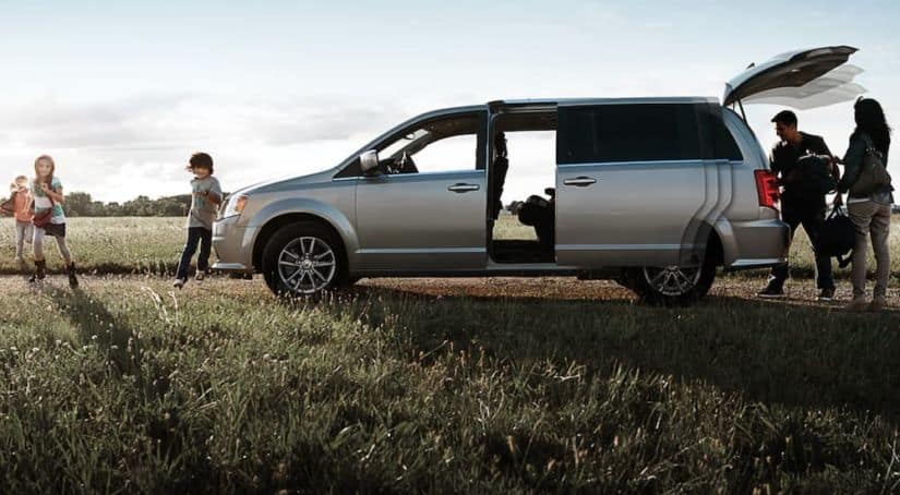 A family is surrounding their silver 2019 Dodge Grand Caravan while in a field.