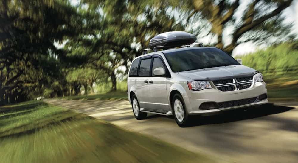 A silver 2019 Dodge Grand Caravan, a popular used car for sale, is driving on a tree-lined road.
