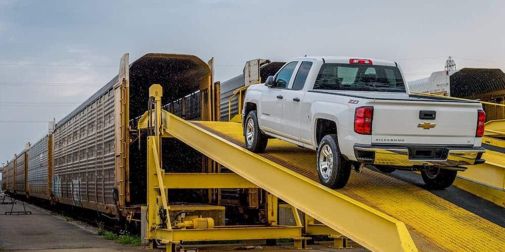 A white 2014 Chevrolet Silverado is being driven up a yellow ramp to be loaded on a train.
