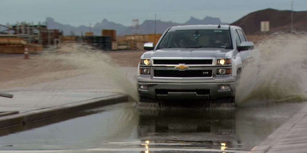 A grey 2014 Chevrolet Silverado is driving through a large puddle splashing water.