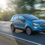 A blue 2016 used Ford EcoSport is driving down the road with the sun shining through the trees.