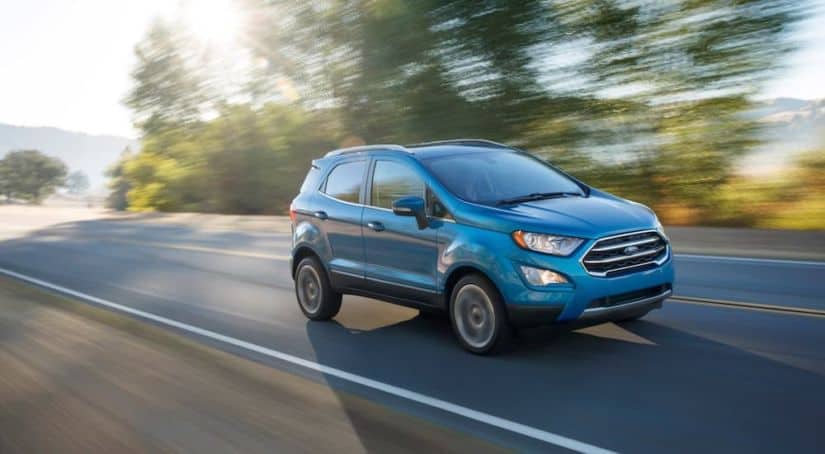 A blue 2016 used Ford EcoSport is driving down the road with the sun shining through the trees.