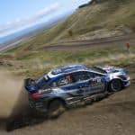 A blue 2018 Subaru WRX STI rally car is drifting up a dirt mountain road kicking up dust.
