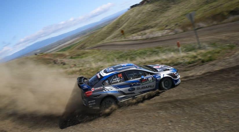A blue 2018 Subaru WRX STI rally car is drifting up a dirt mountain road kicking up dust.
