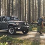 A couple is walking away from a 2020 Jeep Wrangler Unlimited with back packs on a road in the woods.