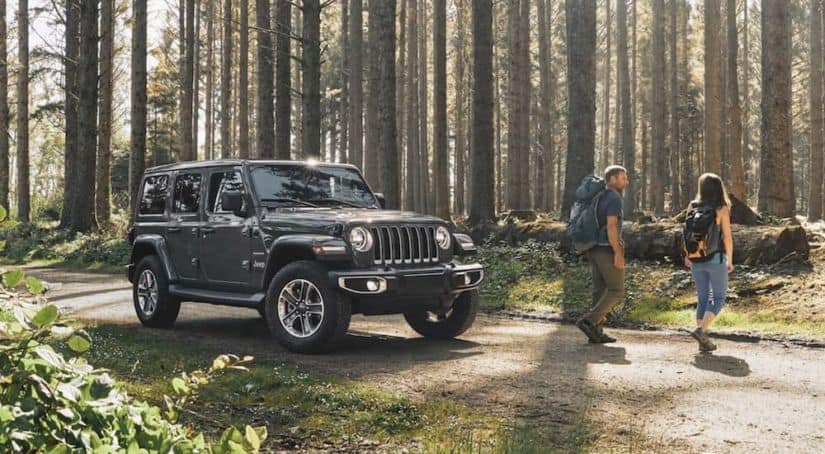 A couple is walking away from a 2020 Jeep Wrangler Unlimited with back packs on a road in the woods.