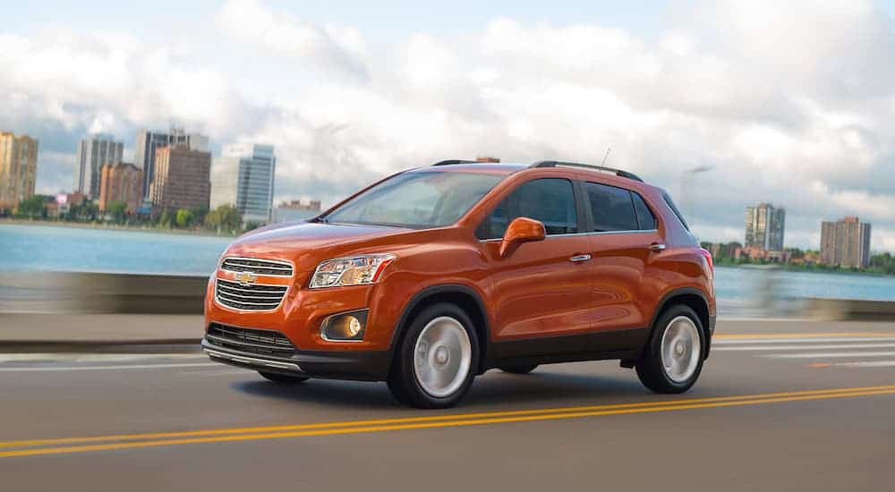An orange 2015 Chevy Trax is driving on a road past a lake and city.