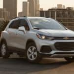 A white 2021 Chevrolet Trax is parked on a city street in front of a city skyline at sunrise.