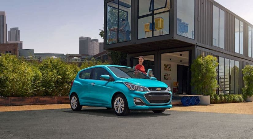 A woman is getting into a teal 2021 Chevy Spark parked in front of a storage unit home.