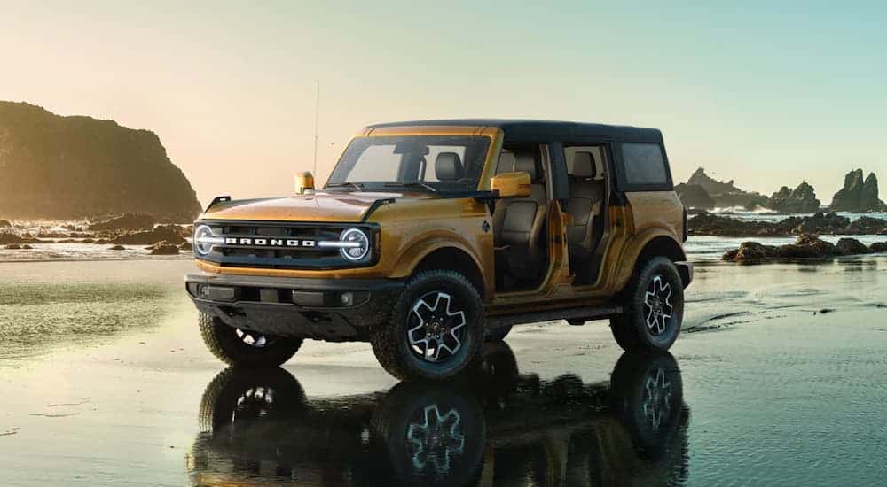 A yellow 2021 Ford Bronco (4dr) is parked on a beach with rock formations in the water after winning the 2021 Ford Bronco (4dr) vs 2020 Jeep Wrangler Unlimited comparison.