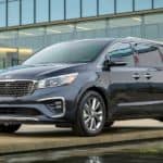 A blue 2021 Kia Sedona is parked in front of a modern building near water.