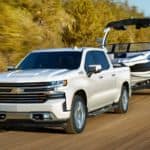 A white 2020 Chevy Silverado 1500 from a Chevy dealer near me is towing a boat past trees.