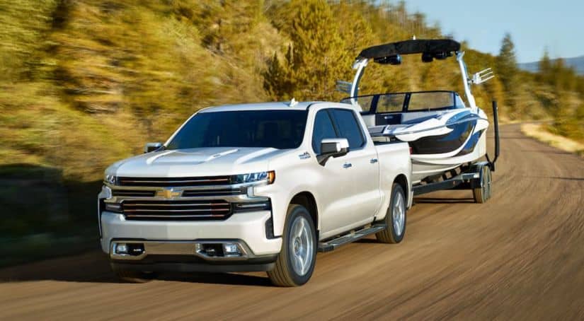 A white 2020 Chevy Silverado 1500 from a Chevy dealer near me is towing a boat past trees.