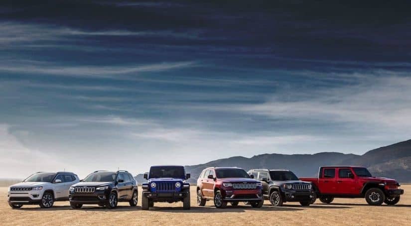A line up of six Jeep models from a Jeep dealership near you are parked in the open in front of mountains.