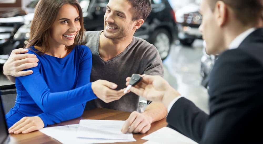 A salesman is handing a couple the keys to a used car with paperwork on the table.