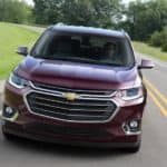 A maroon 2018 used Chevy Traverse is driving on a grass-lined road, shown from the front.