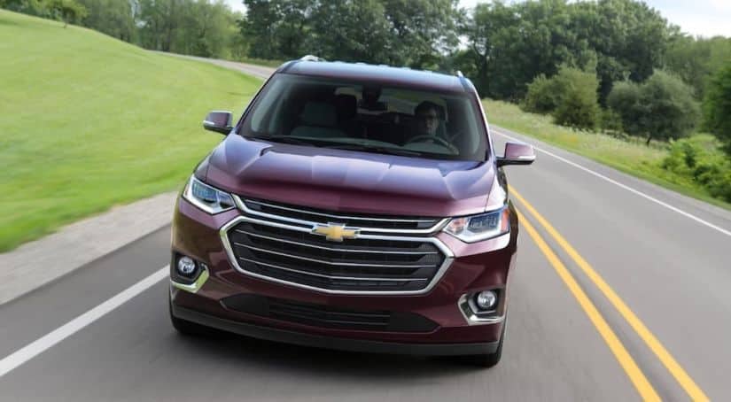 A maroon 2018 used Chevy Traverse is driving on a grass-lined road, shown from the front.
