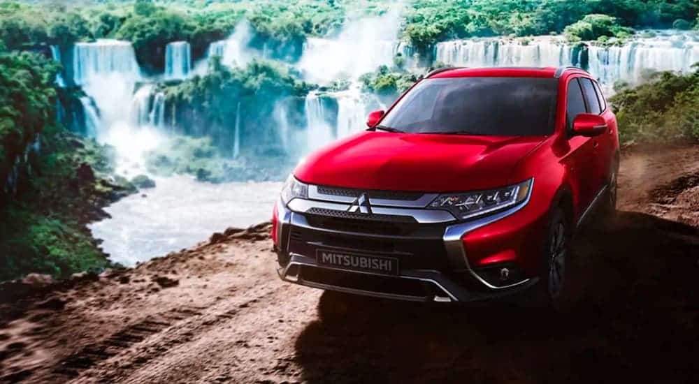 A red 2020 Mitsubishi Outlander is driving on a dirt trail in front of waterfalls.