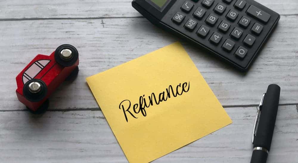 A yellow sticky note that says refinance is sitting on a desk with a pen, calculator, and red toy car.