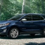 A blue 2020 Chevy Equinox is parked on a gravel driveway at a home in the woods.