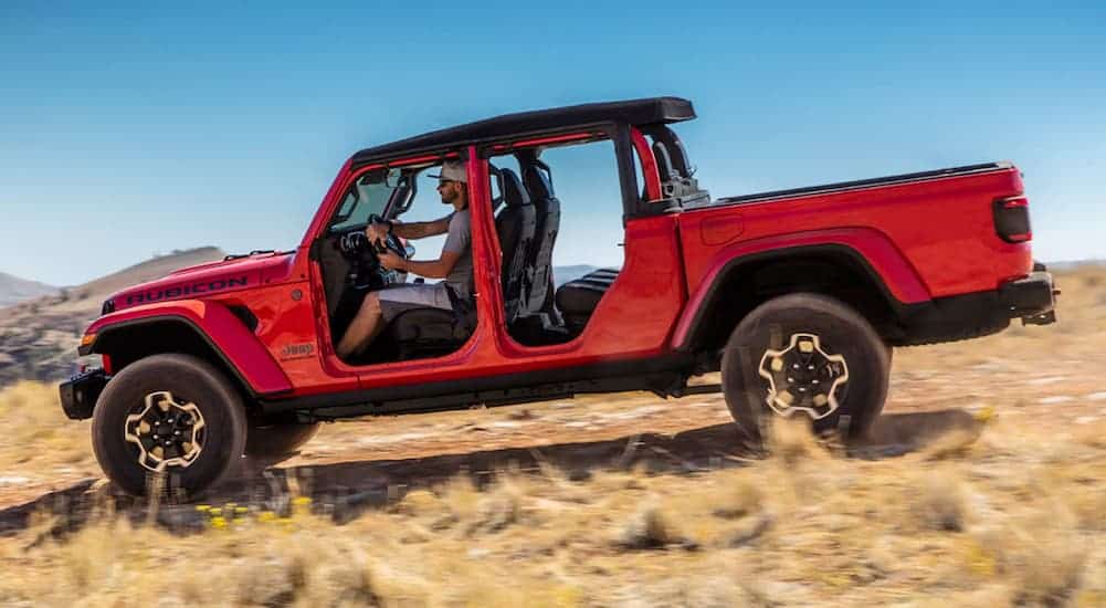 A red 2021 Jeep Gladiator Rubicon is driving on a dirt path past dry grass, shown from the side with no doors.