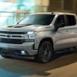 A silver 2021 Chevy Silverado 1500 is driving past blurred buildings with light reflecting off its glossy paint.