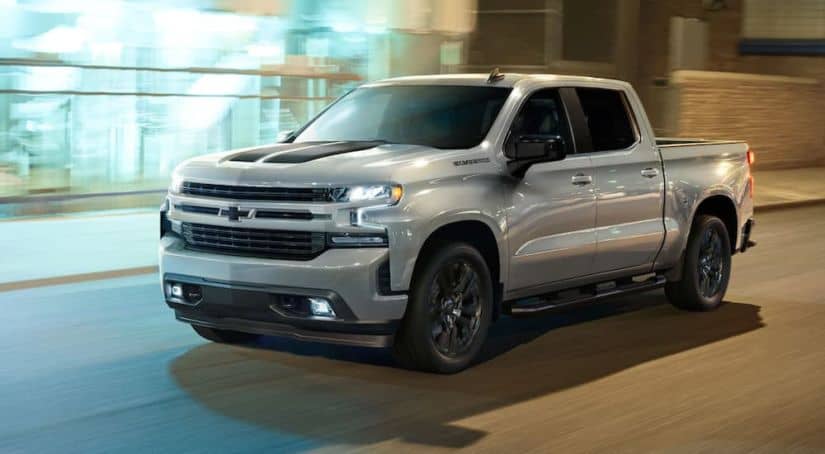 A silver 2021 Chevy Silverado 1500 is driving past blurred buildings with light reflecting off its glossy paint.