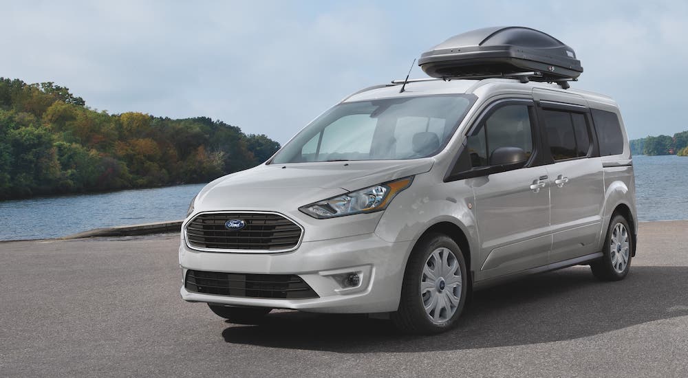A silver 2021 Ford Transit Connect with roof cargo is parked in front of a lake.