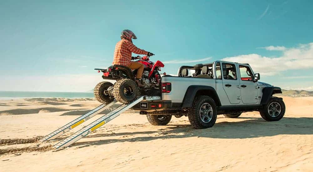 A silver 2021 Jeep Gladiator Rubicon is parked in the desert with an ATV being driven up ramps into the bed.