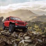 A red 2021 Jeep Wrangler Rubicon Unlimited is parked on a rocky hill with misty mountains in the background.