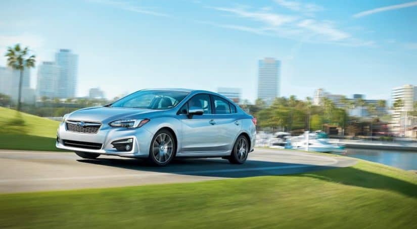 A popular affordable used car, a silver 2017 Subaru Impreza, is driving down the street away from a city and marina.